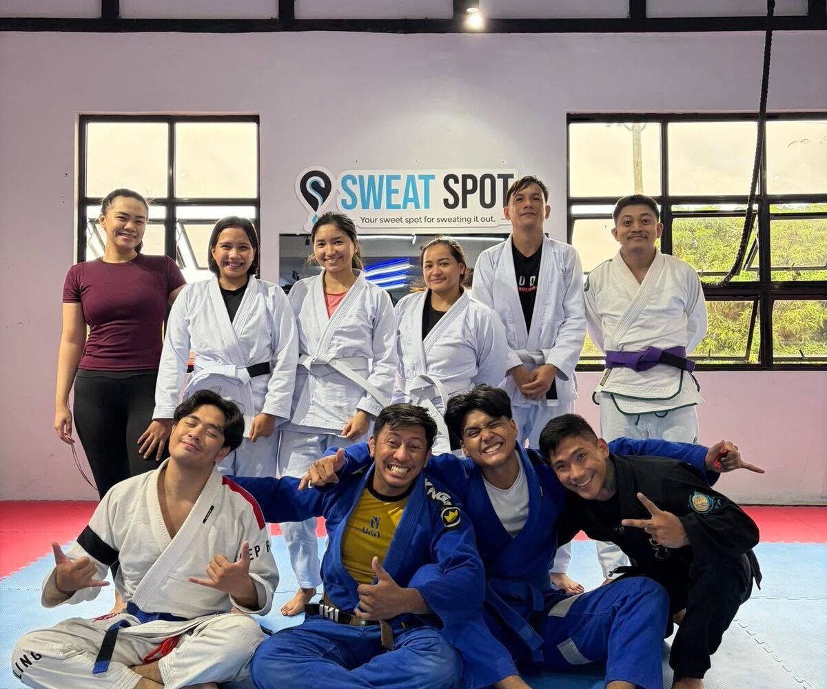 Group of members, most in jiu-jitsu gis, posing together on the mats under the Sweat Spot sign