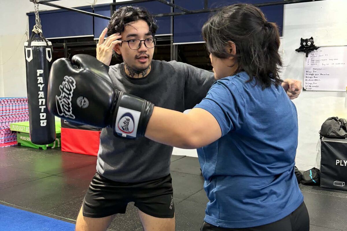 A coach in a grey tee guiding a member in a blue tee through a punch in black boxing gloves, beside a heavy bag on the gym floor