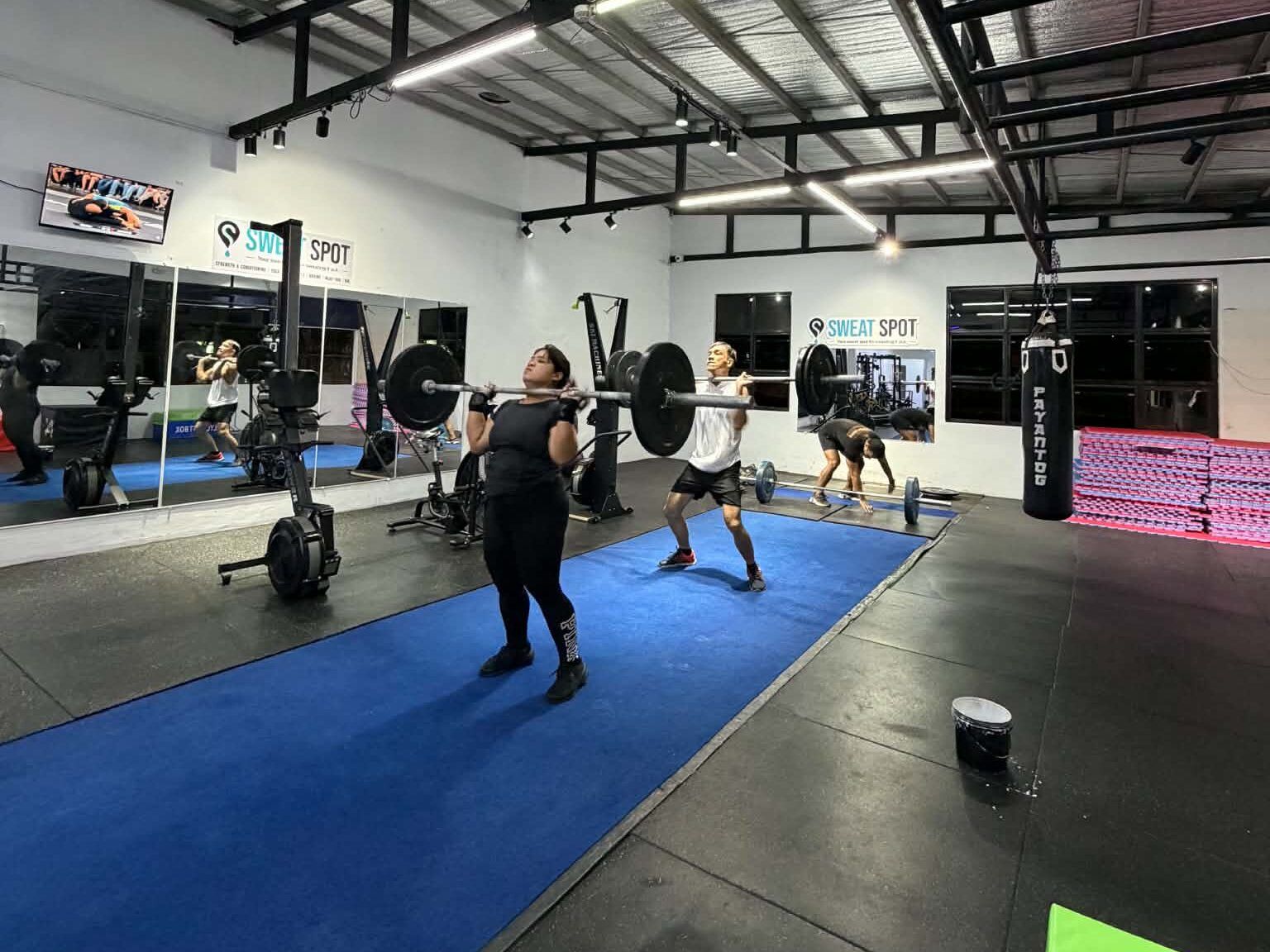 Members lifting barbells on blue turf in the Sweat Spot gym, with the Sweat Spot sign on the wall behind them